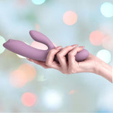 A hand holding the Svakom Trysta Neo Rabbit Vibrator in Romantic Rose, a dual-motor personal massager designed for G-spot stimulation, against a background of soft, out-of-focus pastel lights.